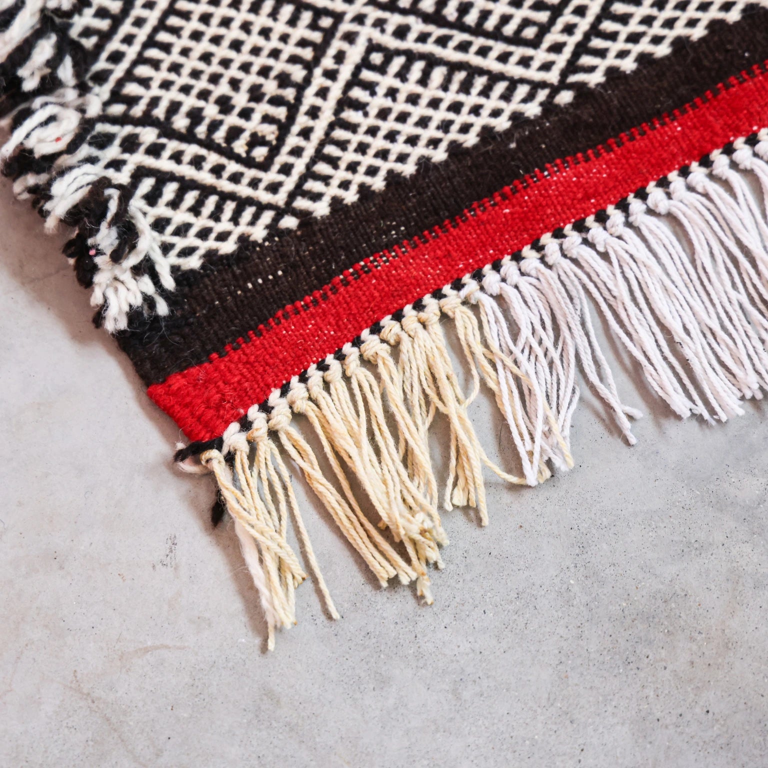 Close-up of a woven textile with geometric pattern and fringes on a concrete surface