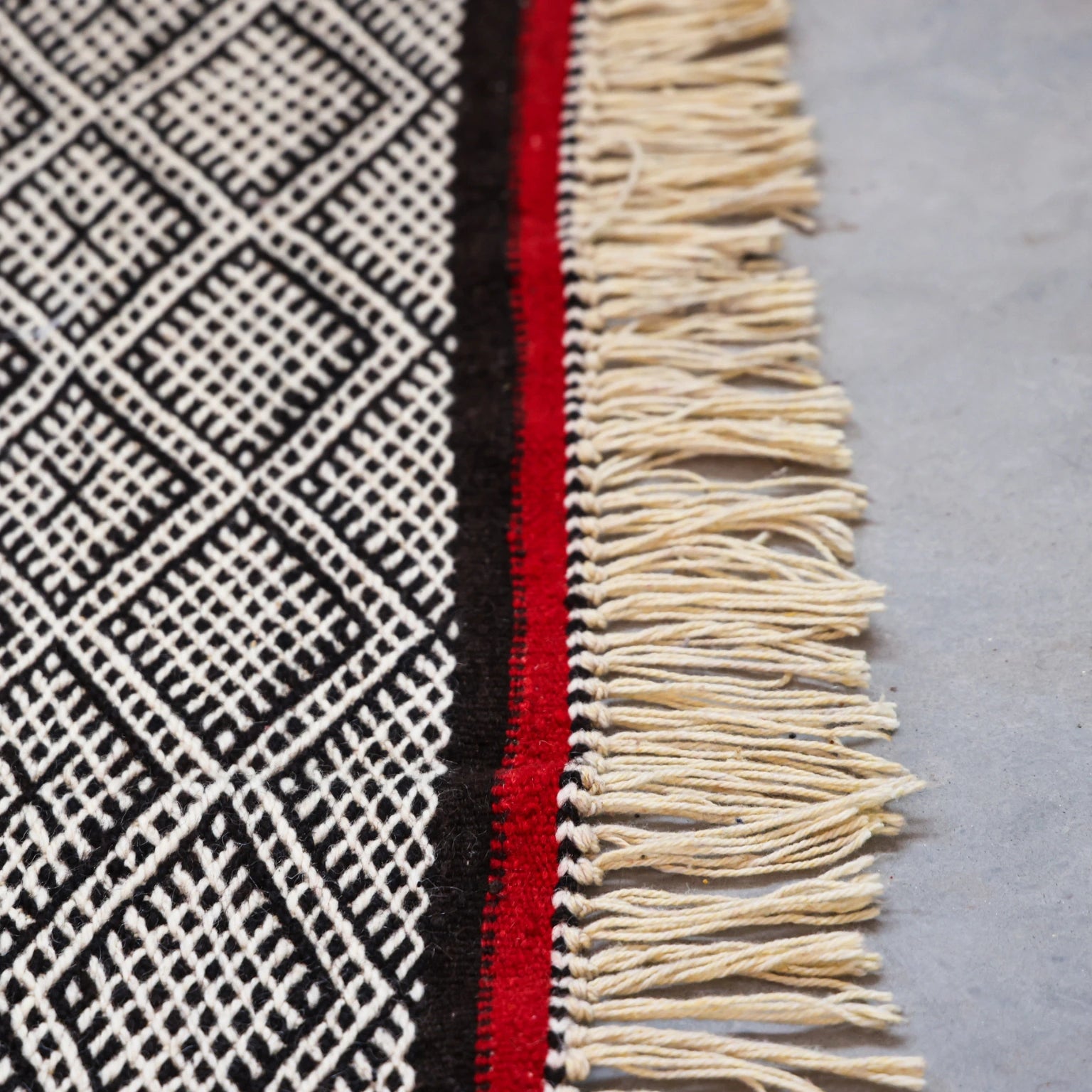 Textile with geometric pattern, red stripe, and beige fringe on a gray surface